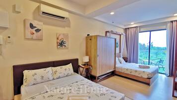 Quarto de hotel com duas camas, um guarda-roupa, uma mesa de cabeceira com abajur e vista para o verde pela janela.