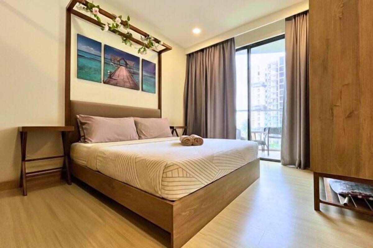Une chambre d’hôtel moderne avec un grand lit, une tête de lit en bois, une petite table et une vue sur la ville grâce à de grandes fenêtres.
