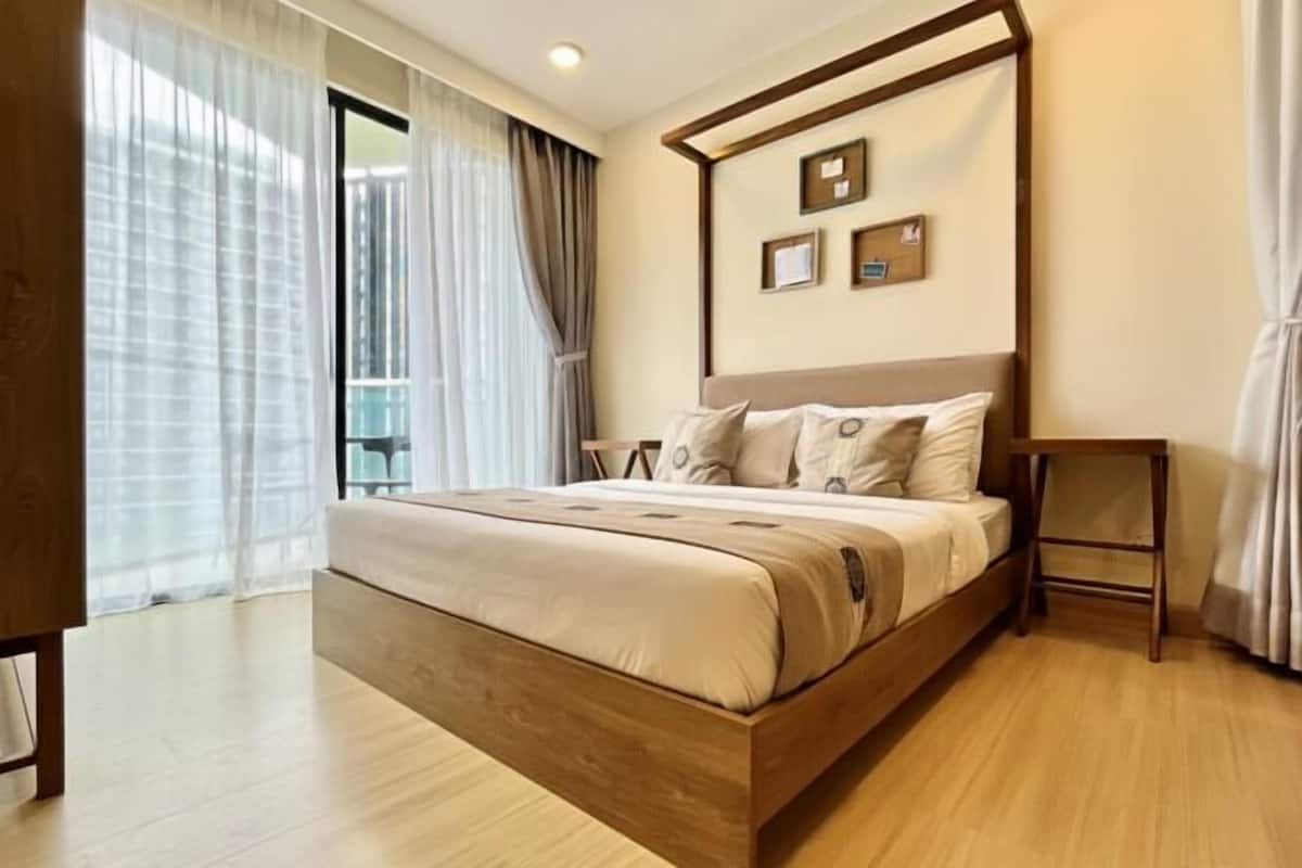 A modern bedroom with a wooden bed, a bedside table, and a view of the outdoors through sheer curtains.