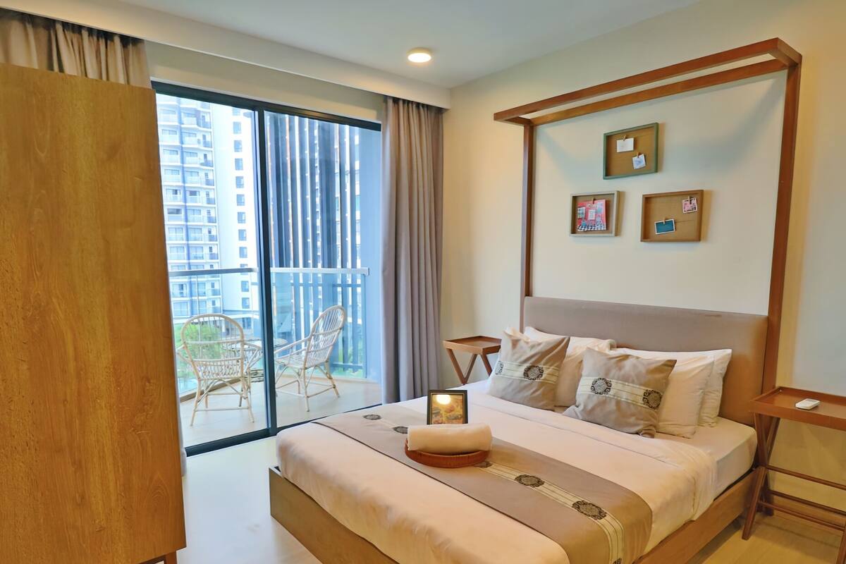A hotel room with a large bed, a bedside table, a nightstand, a window with a view of buildings, and a balcony with chairs.