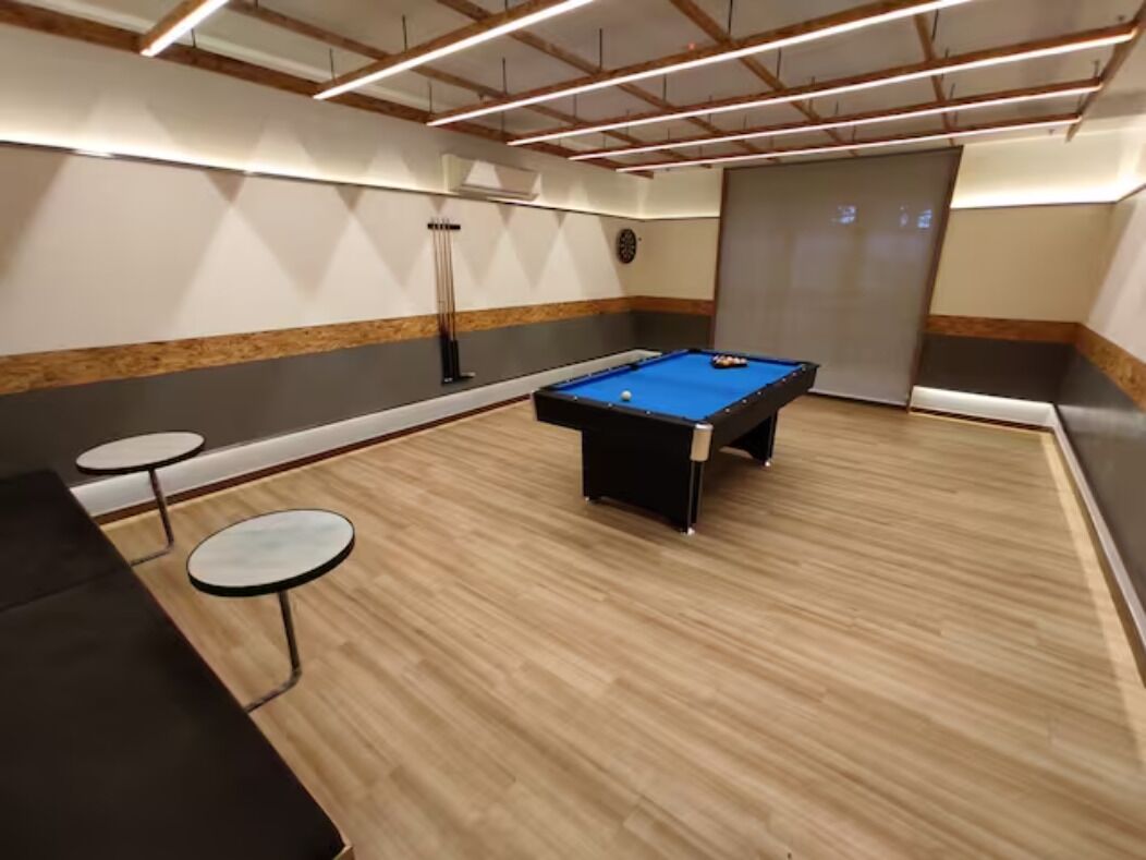 Mesa de bilhar em um espaço com piso de madeira, duas pequenas mesas redondas e design moderno no teto.