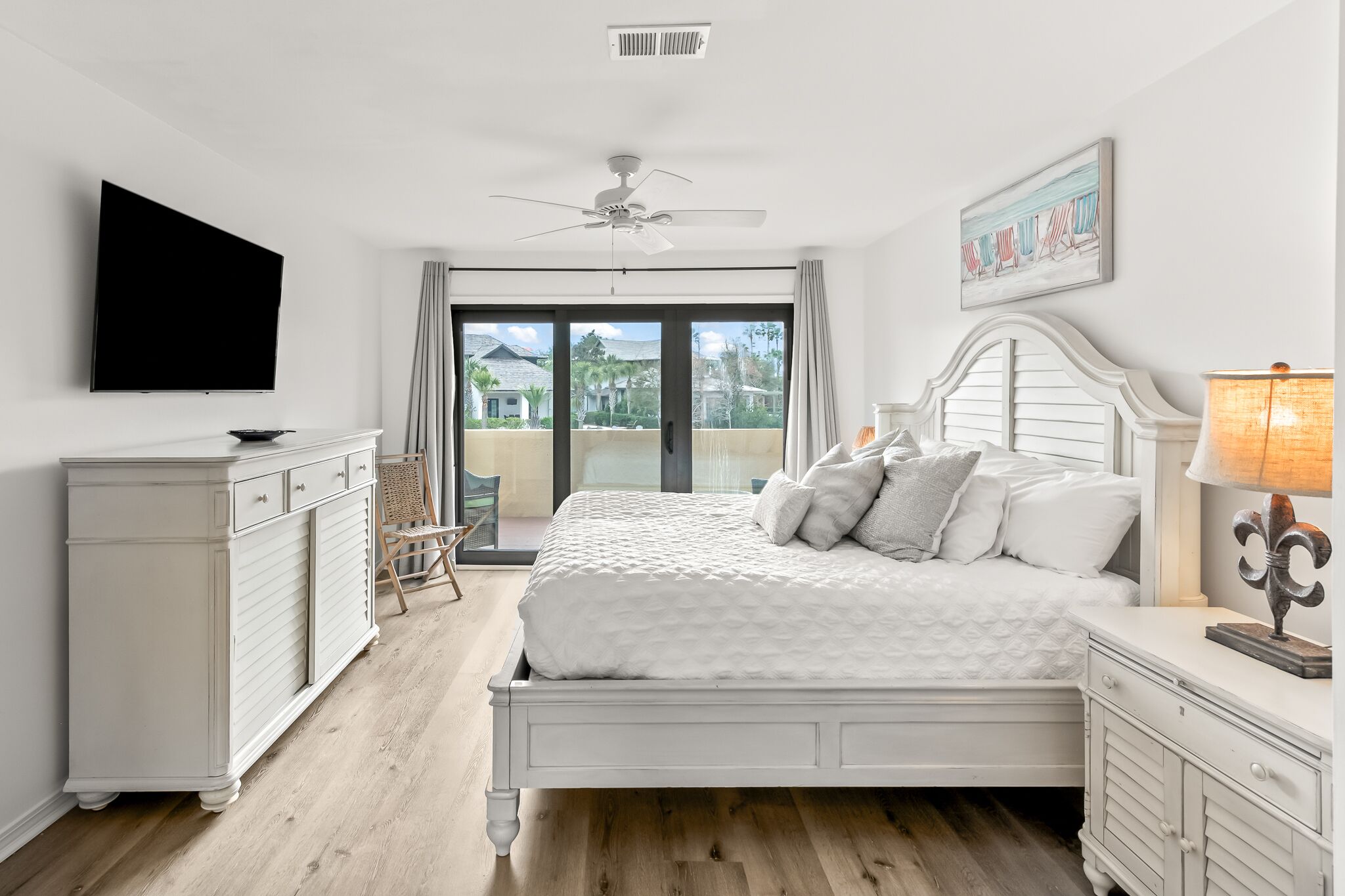 A bedroom with a bed, a dresser, a television, a ceiling fan, and a balcony with a view of houses and palm trees.