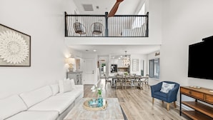Een ruime woonkamer met een witte bank, een salontafel en een blauwe fauteuil. Een trap leidt naar een hoger niveau met een balustrade.