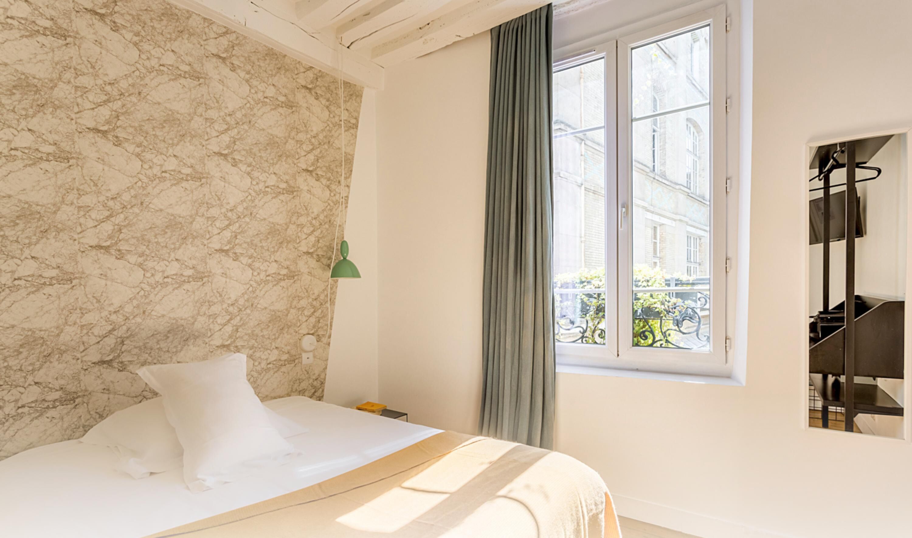 Une chambre avec un mur en marbre, un lit recouvert de draps blancs, une lampe murale verte et une fenêtre donnant sur des immeubles.