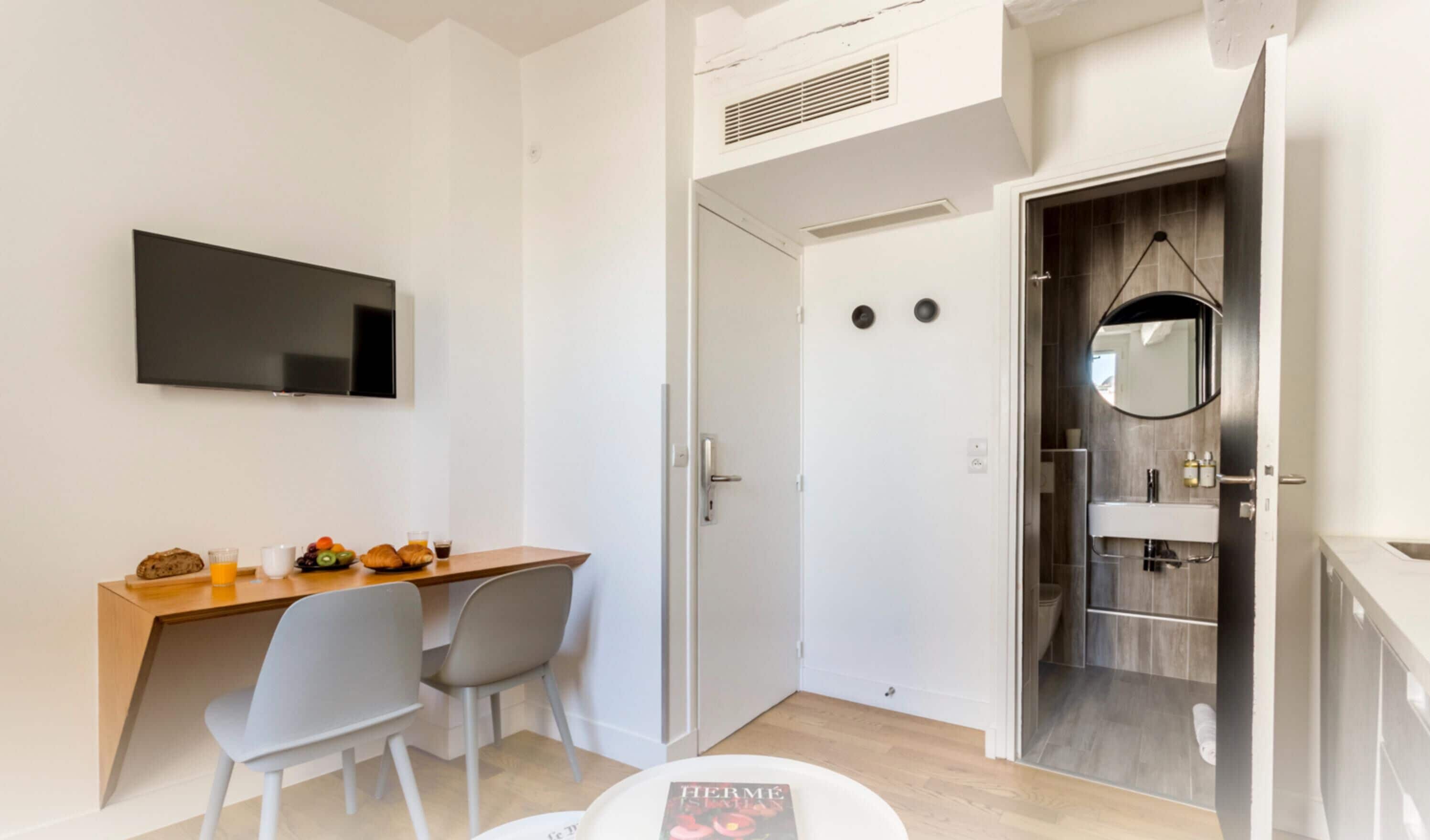 Een moderne hotelkamer met een houten bureau, twee stoelen, een flatscreen televisie, een badkamer met spiegel en wastafel, en een deur naar een andere kamer.