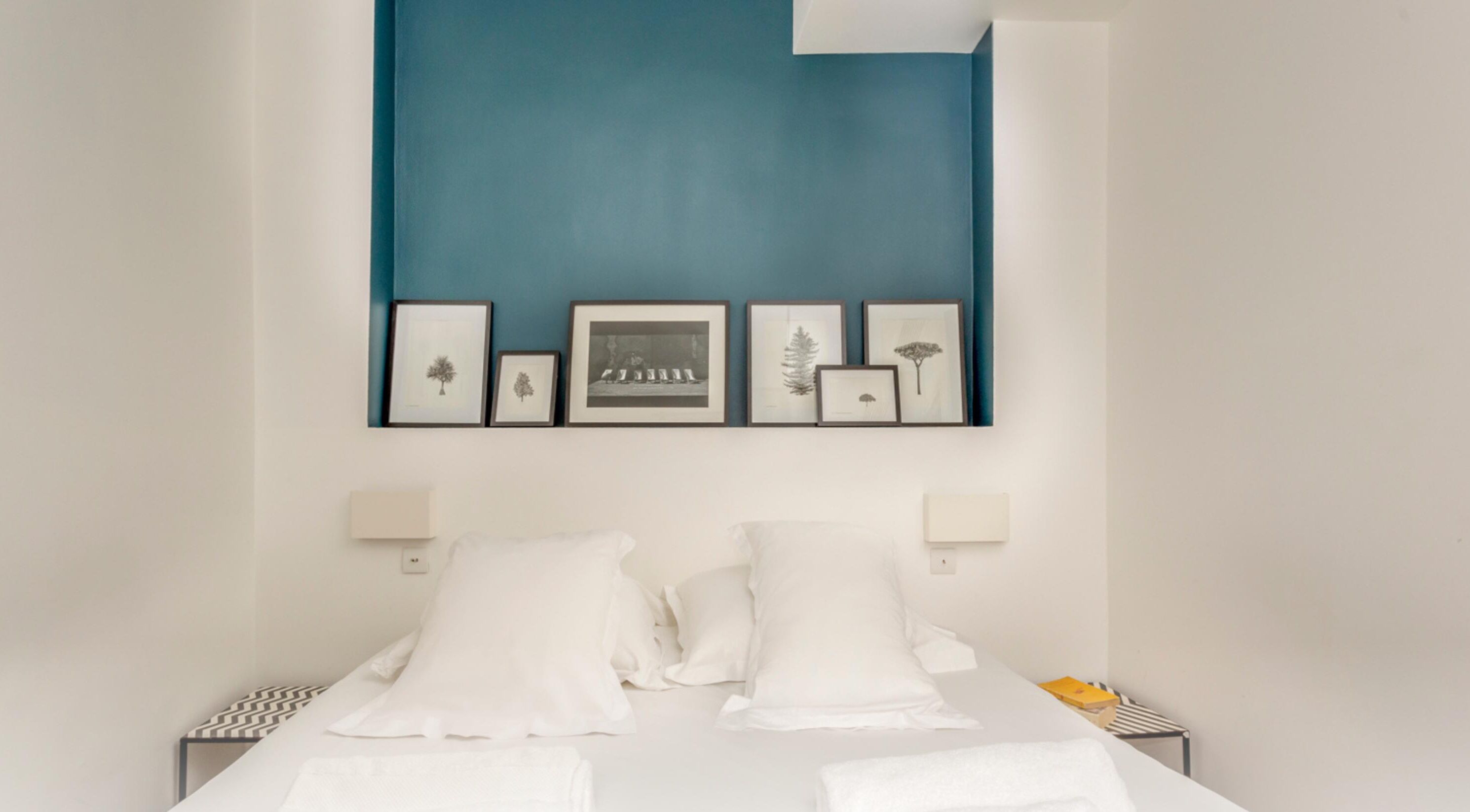 Un dormitorio con cama, almohadas blancas, una pared de acento azul y cuadros enmarcados.