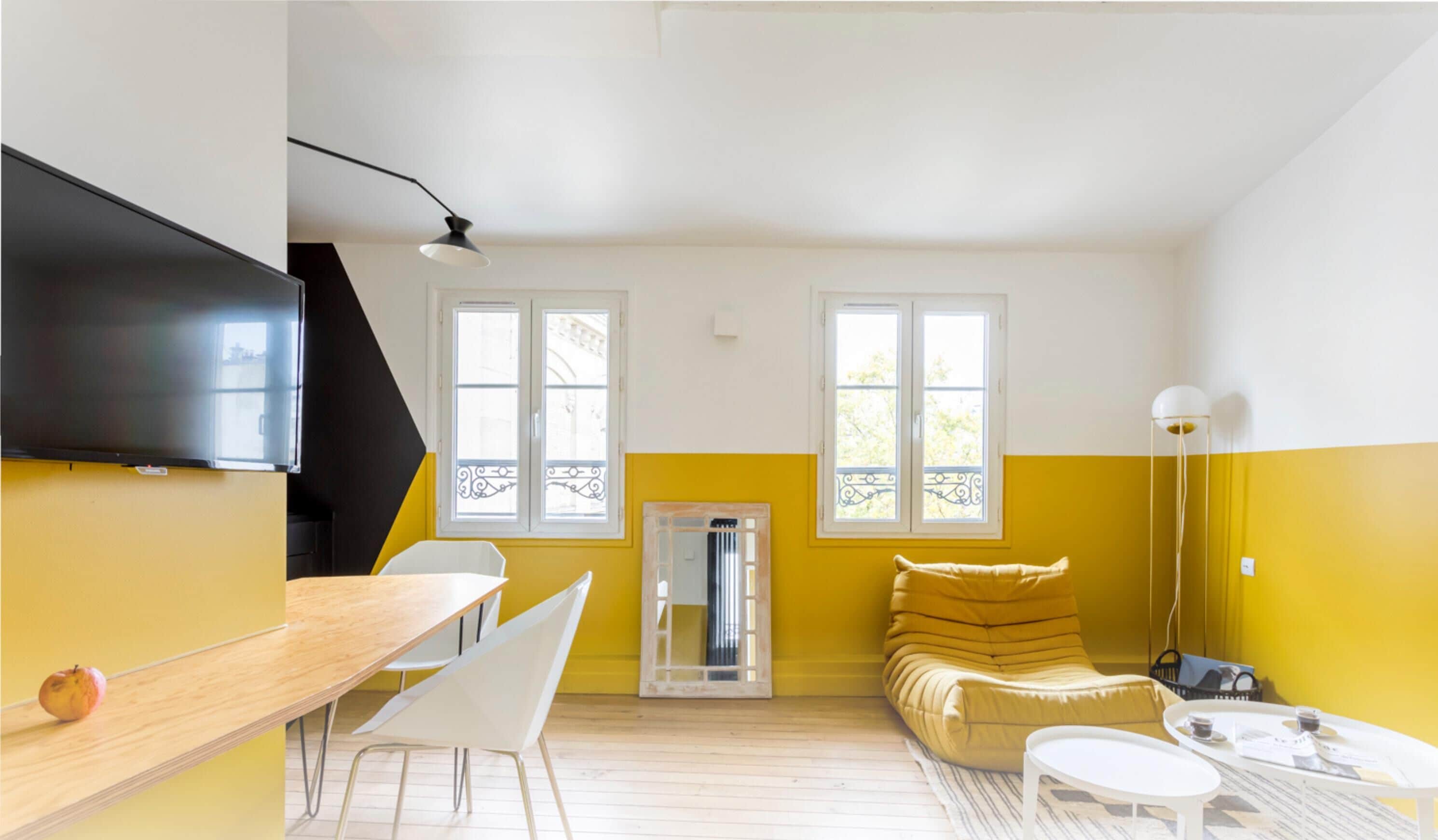 Ein modernes Wohnzimmer mit einem Holzesstisch, weißen Stühlen, einem gelben Sofa und einem an der Wand befestigten Fernseher.