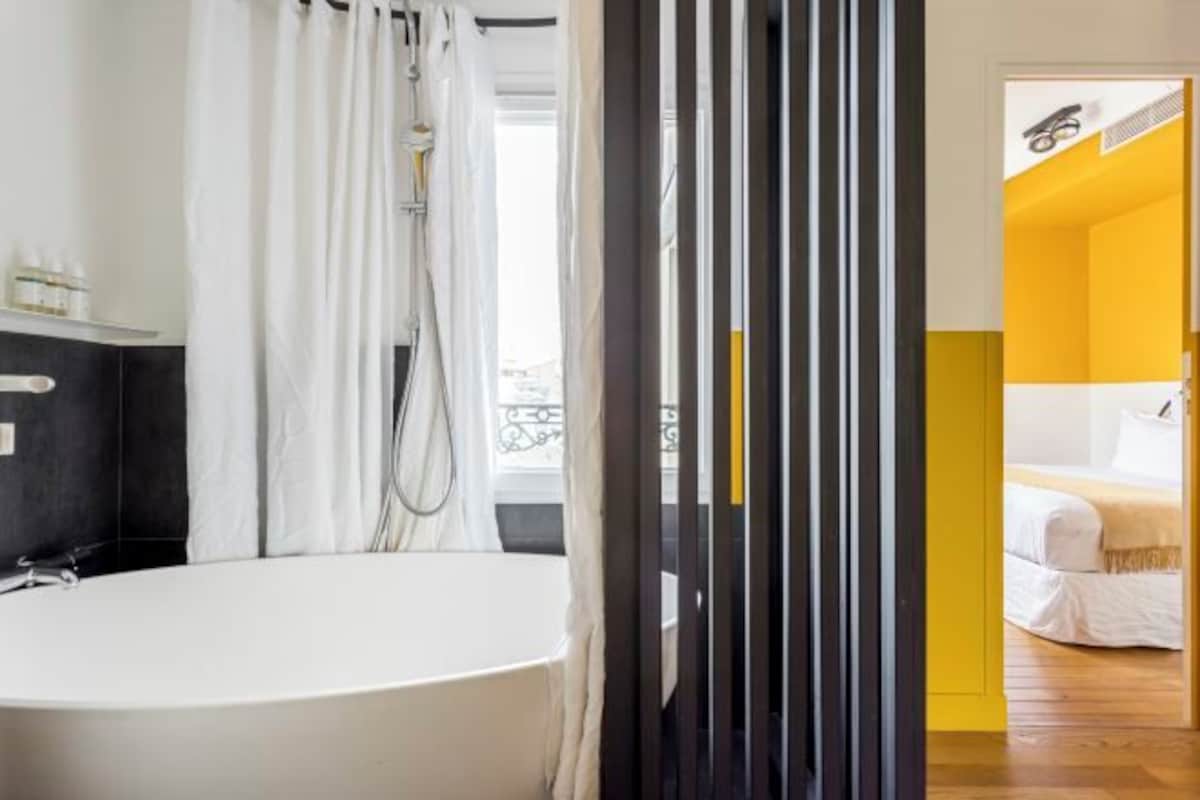 Un baño moderno con bañera, cortina de ducha blanca y vista a un dormitorio con cama y pared amarilla.