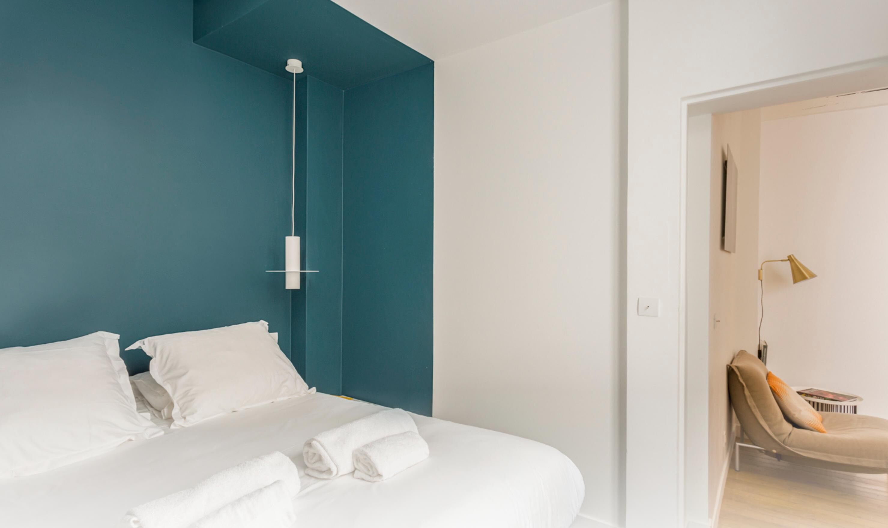 A modern hotel room with a teal accent wall, a bed with white linens, and a single roll of white towels.