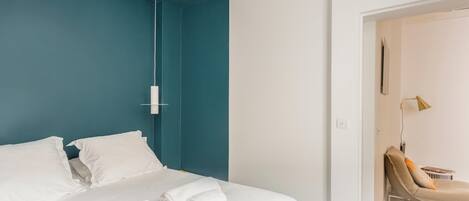 A modern hotel room with a teal accent wall, a bed with white linens, and a single roll of white towels.