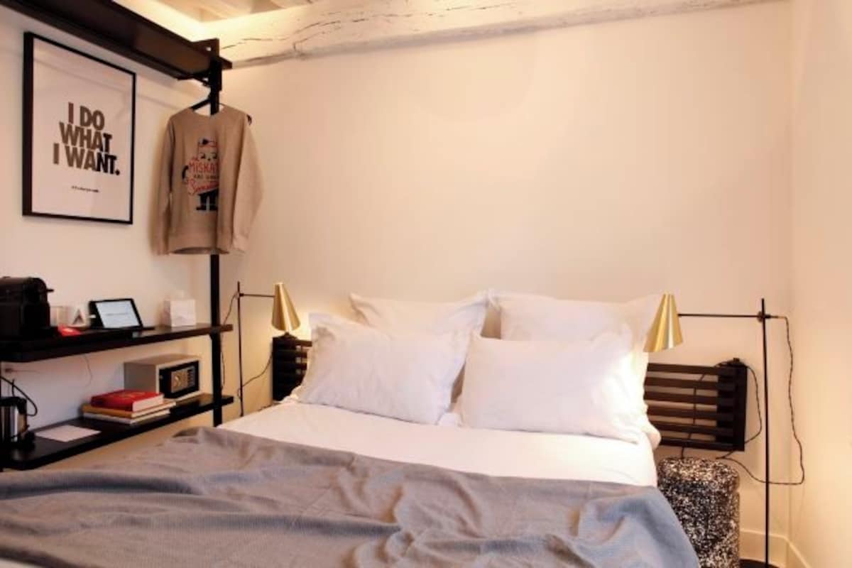 Una cama bien tendida con sábanas blancas, una mesita de noche con lámpara y un cartel enmarcado en la pared.