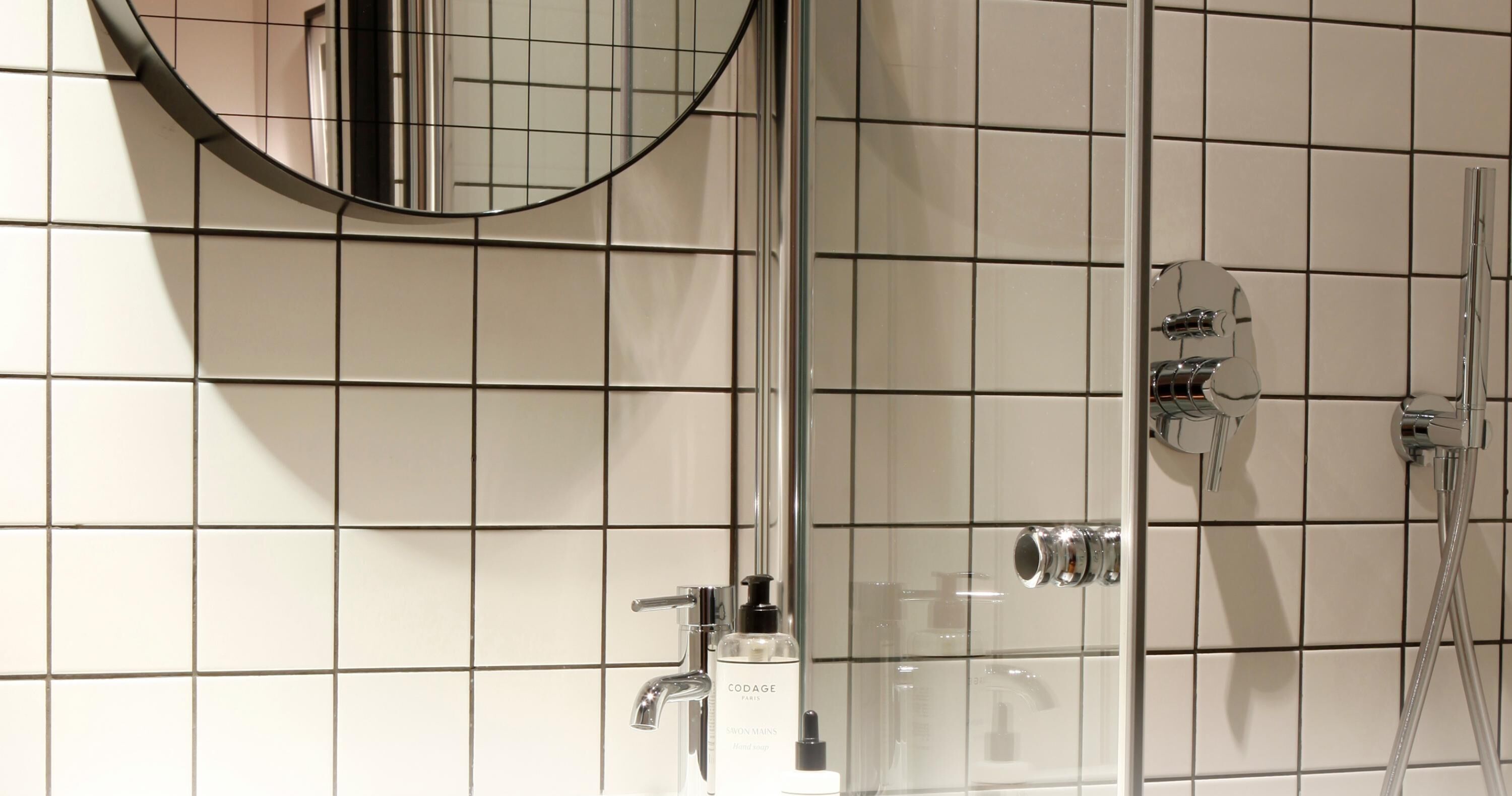 Une salle de bain avec un mur carrelé, un miroir rond, une cabine de douche en verre et un distributeur de savon.
