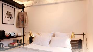 Camera d'albergo ordinata con un letto, cuscini bianchi, un comodino con una lampada, una citazione motivazionale incorniciata e una felpa appesa.