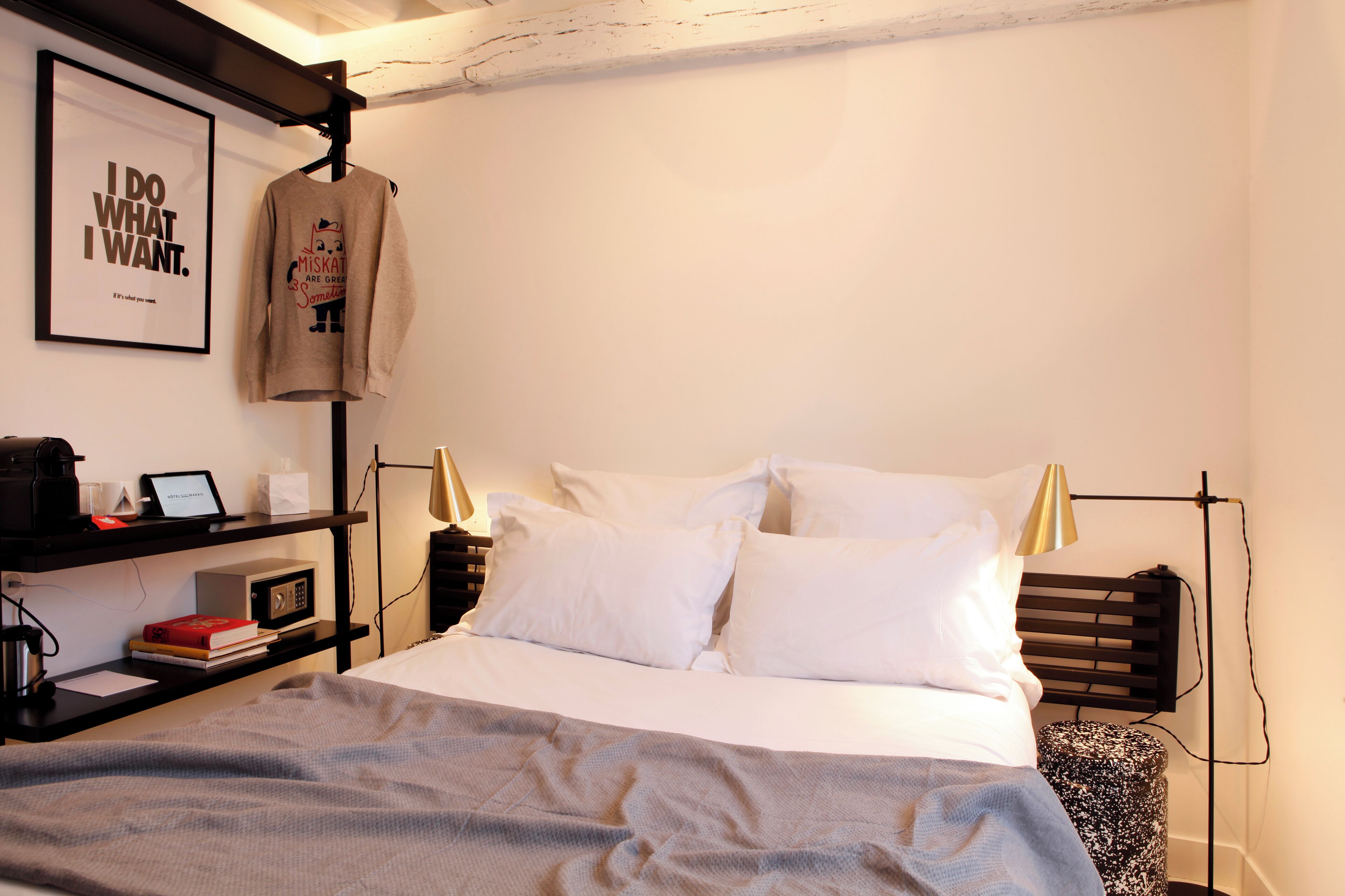 A neatly made bed with white linens, a grey blanket, and a wall-mounted shelf with books and a coffee machine.