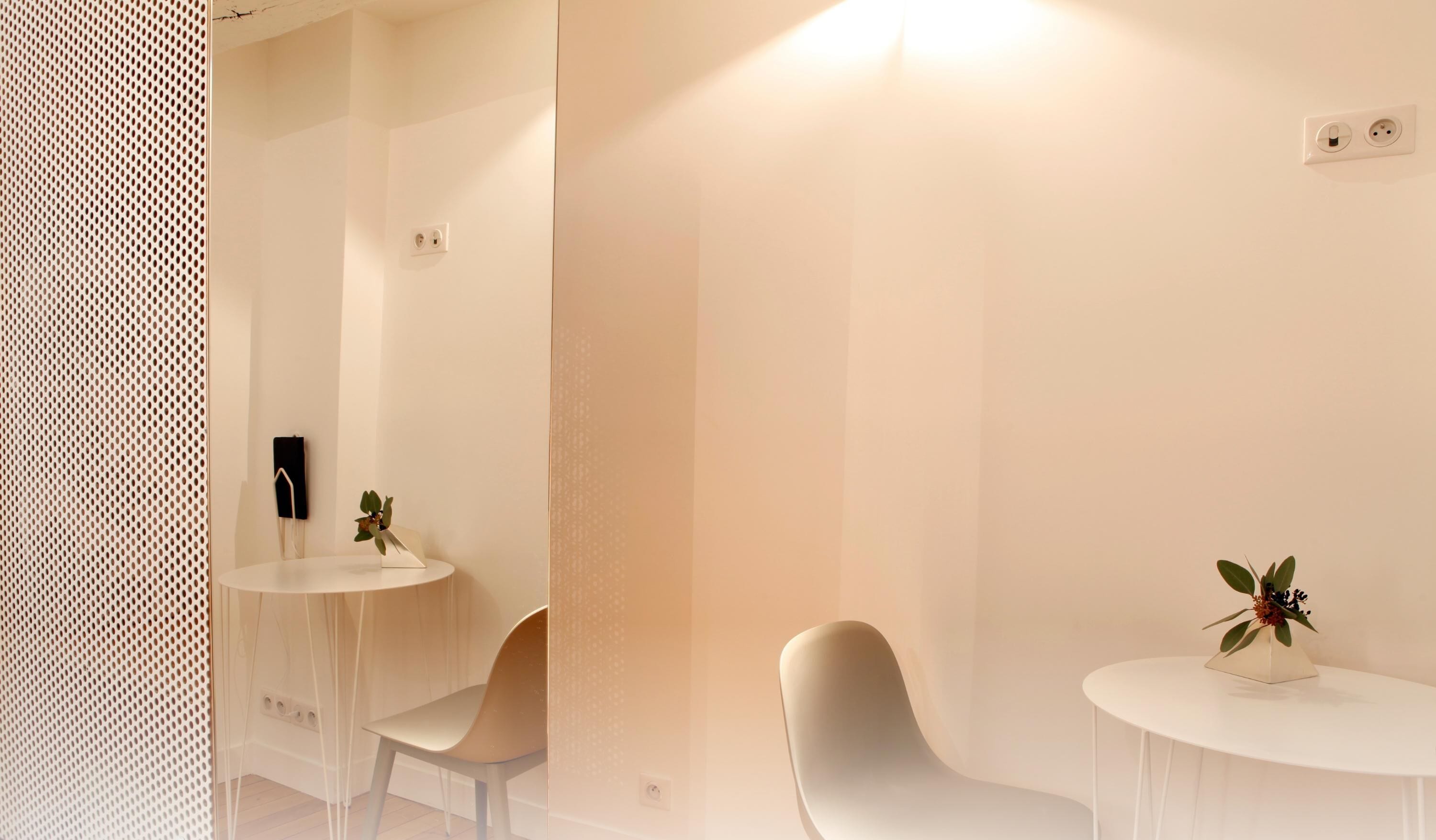 A modern, minimalist room with white walls and furniture. There are two white tables, each with a potted plant. The room has a clean, uncluttered design with a perforated wall on the left.