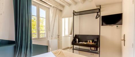 A bedroom with a bed, a TV mounted on the wall, a desk with a coffee maker, and a window with curtains.