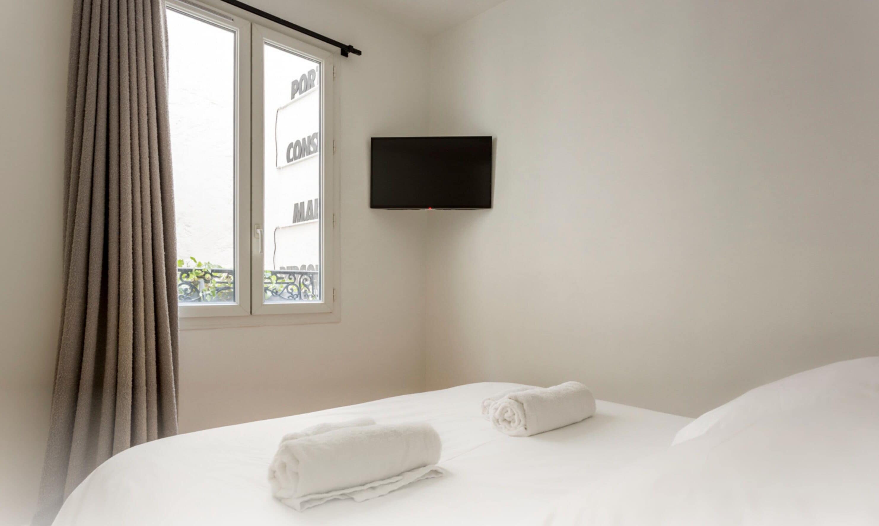 Habitación de hotel con cama, dos toallas enrolladas, ventana con cortinas y televisor montado.