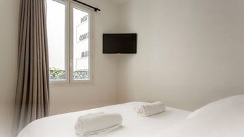 Habitación de hotel con cama, dos toallas enrolladas, ventana con cortinas y televisor montado.