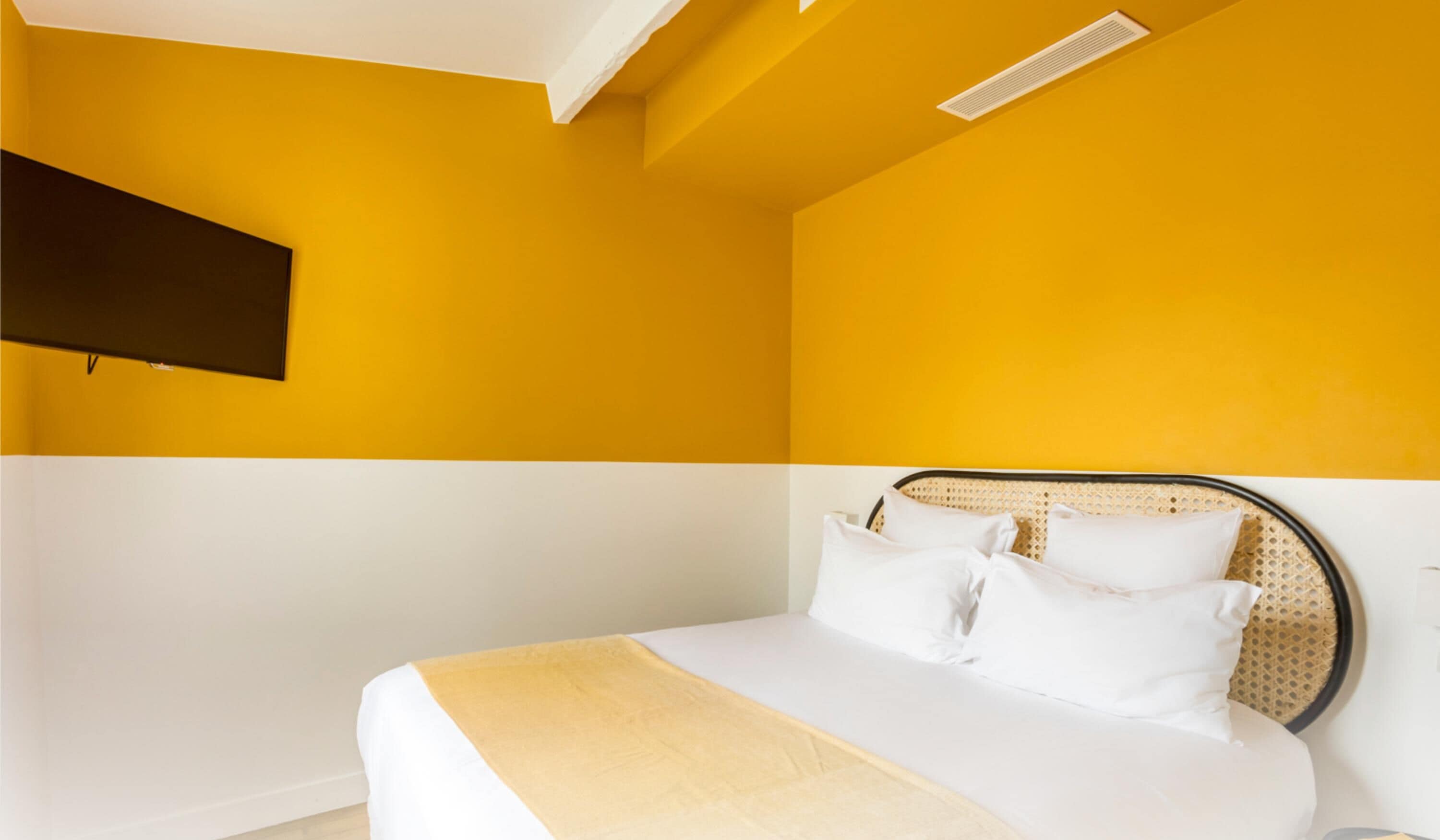 Une chambre à coucher avec un lit, du linge de lit blanc, un mur d’accent jaune et un téléviseur fixé au mur.