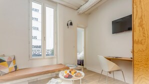 Una habitación moderna y luminosa con un ventanal grande, un escritorio de madera, una silla blanca, una mesita redonda con desayuno y un televisor montado en la pared.