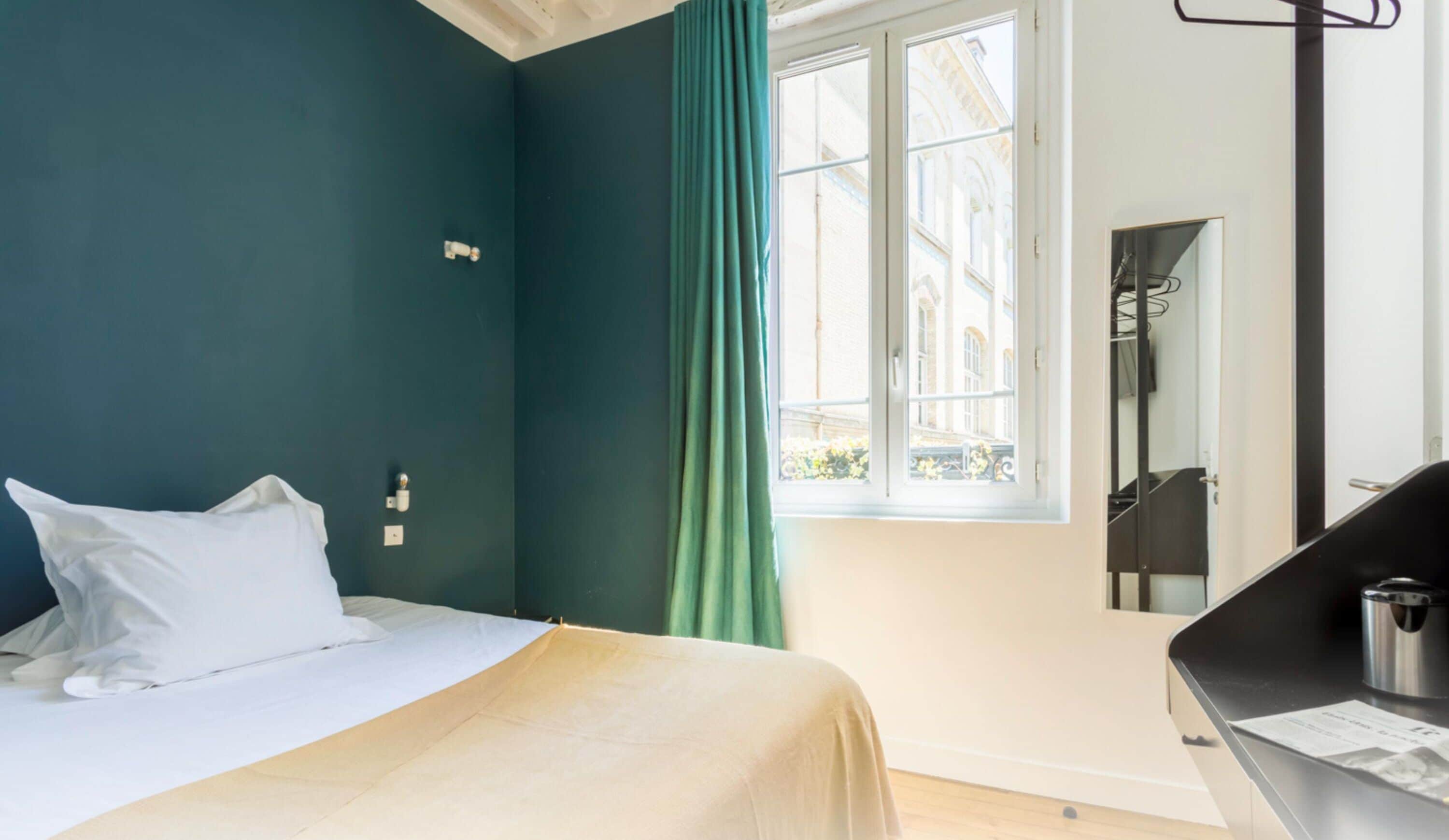 A hotel room with a bed, a window with green curtains, a mirror, and a black desk with a kettle.