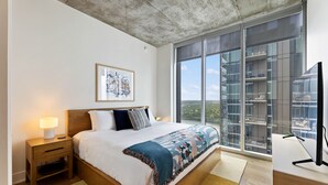 A modern bedroom with a large bed, a nightstand, a bedside lamp, a framed artwork, and a large window offering a view of the city. - Sunshine Condo in Downtown Austin (Austin)