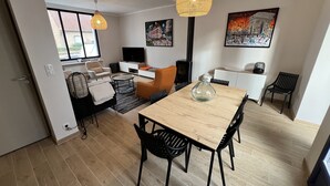 A modern living room with a wooden dining table, black chairs, a sofa, and a TV. There are two hanging lights and a painting on the wall.
