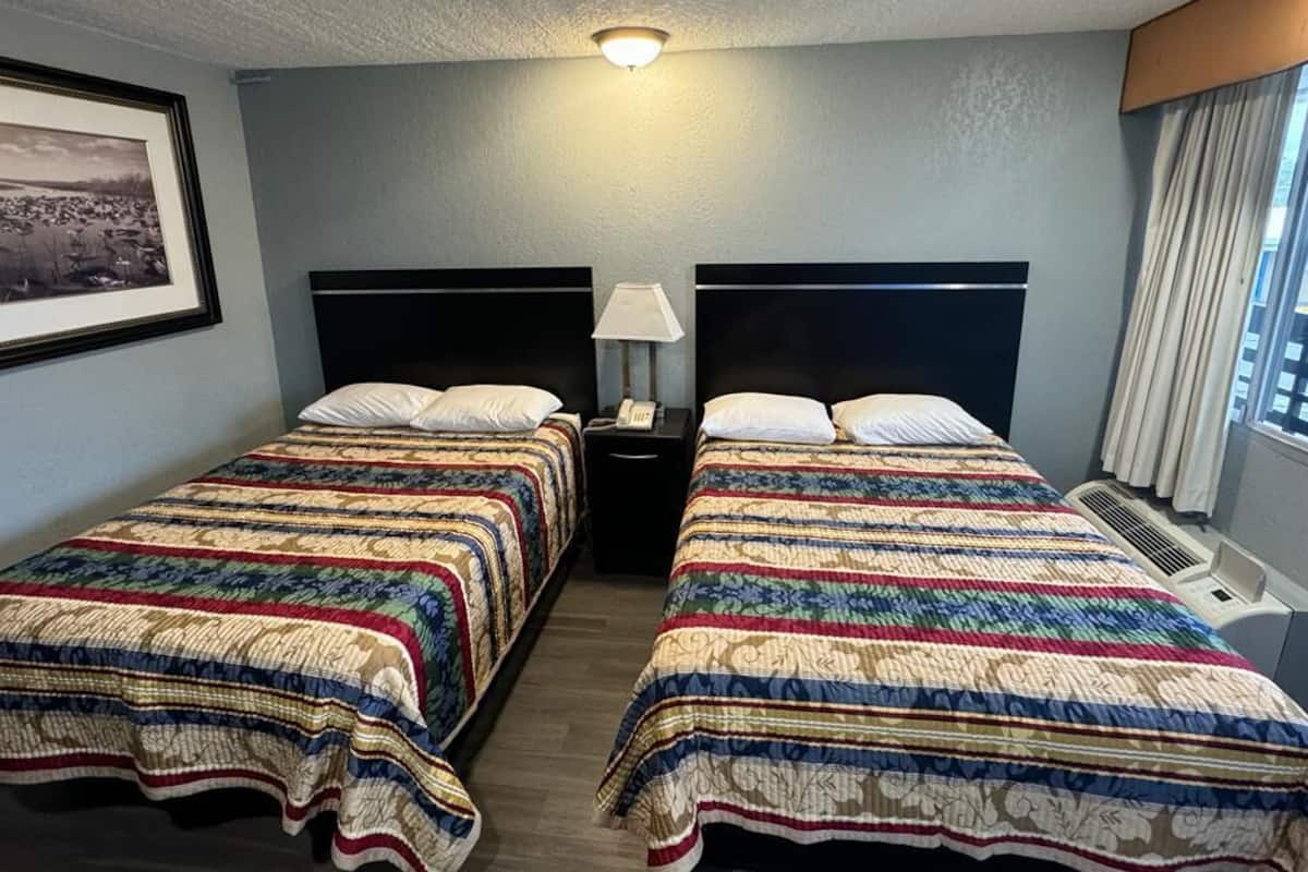 A hotel room with two beds, a nightstand with a lamp, a framed picture on the wall, and a window with curtains.
