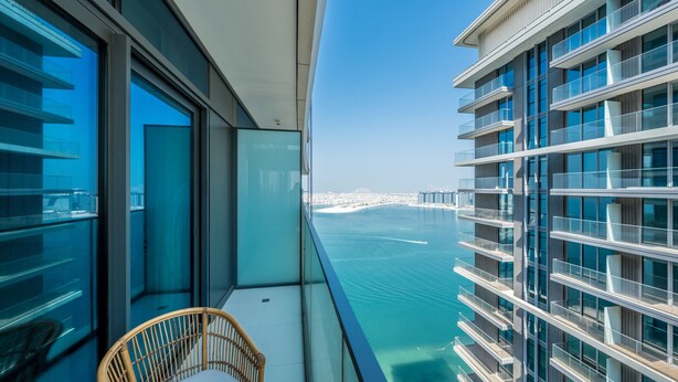 A balcony with a wicker chair, overlooking a body of water and a distant cityscape. - Tanin - Exquisite 1BR Apartment with Sea View  (Dubai)
