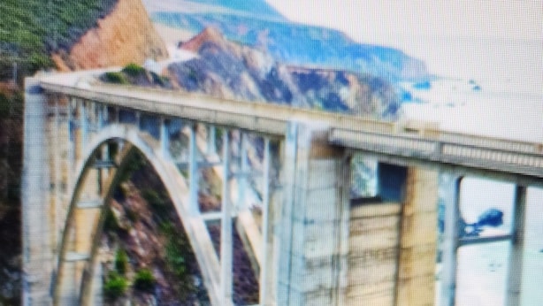 A bridge with a stone arch and a metal structure, surrounded by greenery and mountains in the background.