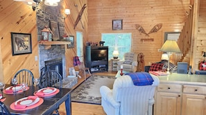 L’intérieur chaleureux d’un chalet avec une cheminée en pierre, des murs en bois, un téléviseur et un coin repas aménagé pour quatre personnes.