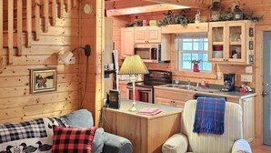 En mysig stuga med en trätrappa, ett köksutrymme, ett vardagsrum med soffa och fåtöljer samt ett matbord.