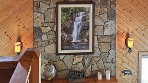 A room with a stone fireplace, a ceiling fan, and a framed picture of a waterfall. - *FAMILY GETAWAY*Deck/Firepit/HOT TUB & Fireplace (Blairsville)