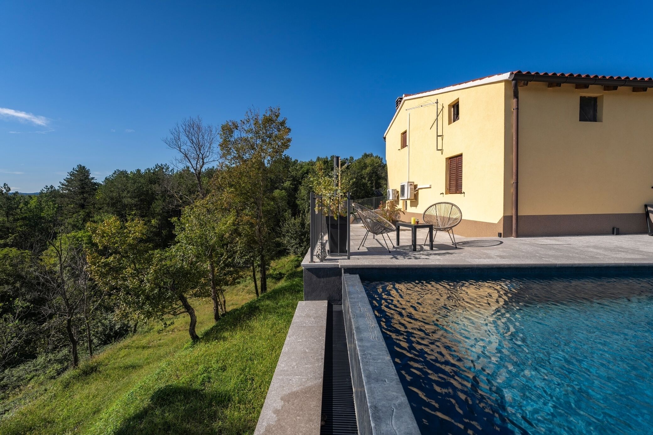 A poolside area with a chair and table, a building with a tiled roof, and a grassy landscape with trees.