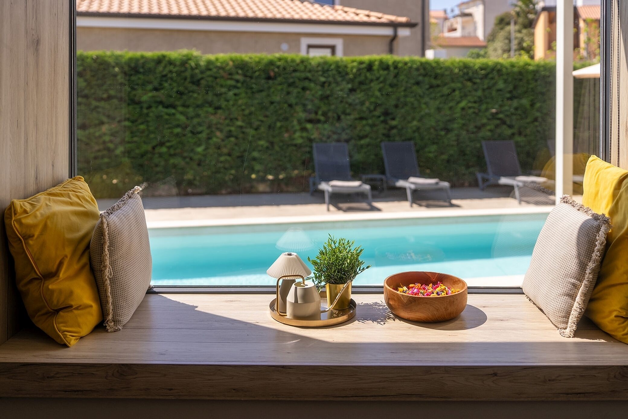 A window seat with a view of a swimming pool, lounge chairs, and a wooden bowl with flowers.