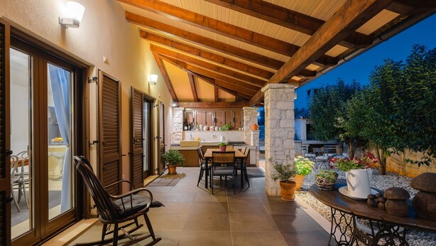 Un espace extérieur aménagé en patio, avec une table à manger, des chaises et un fauteuil à bascule. On y trouve également un pilier en pierre et une table ornée de plantes en pot.