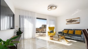 Una sala de estar moderna con sofá, sillón y mesa de centro. Hay un ventanal con cortinas translúcidas, vista a la ciudad y un cuadro en la pared.