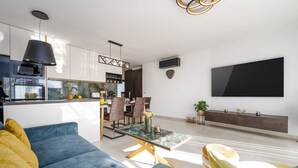 Una sala moderna con un sofá azul, sillones amarillos, una mesa de centro de vidrio y una zona de comedor con sillas, con una cocina al fondo.