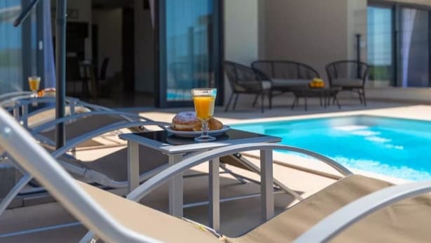 Zona de piscina con sillones de descanso y una mesa con un vaso de zumo de naranja y un pastel.