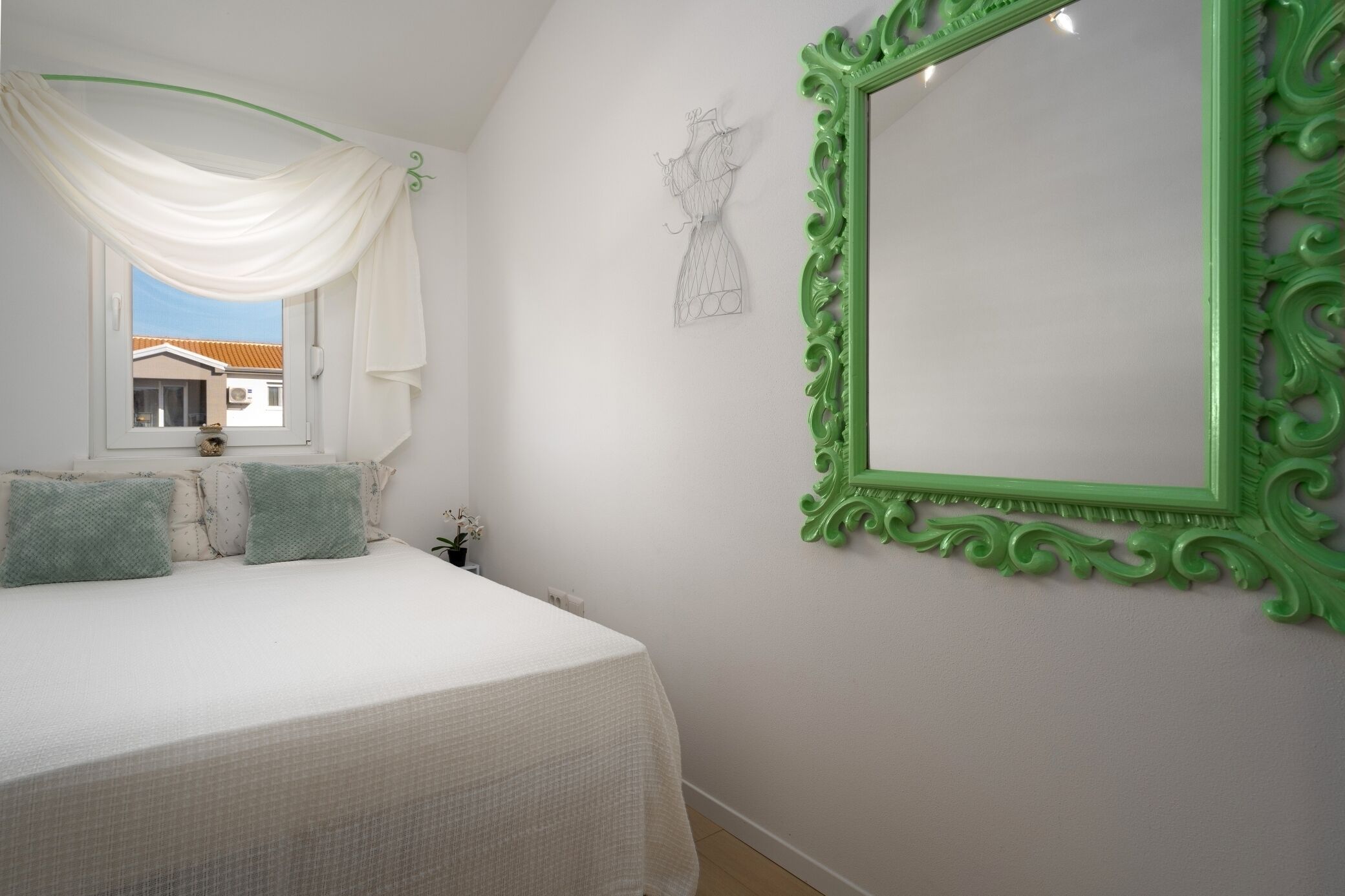 A bedroom with a bed, white curtains, a green-framed mirror, and a wall decoration.