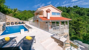 Una casa de dos pisos con piscina, mobiliario exterior y vistas a la vegetación circundante.