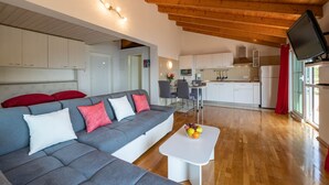 Una sala de estar moderna con un sofá gris, una mesa de centro blanca con fruta, y una cocina al fondo.