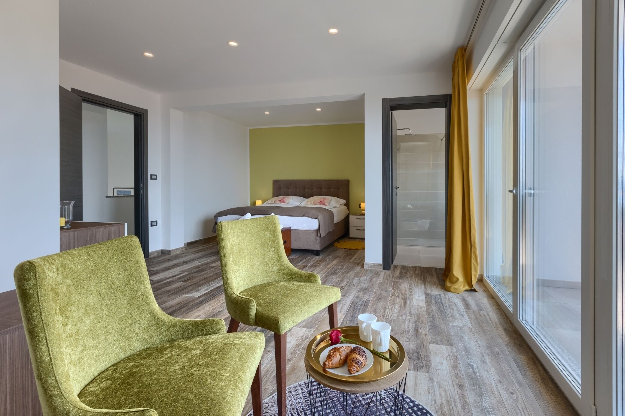 A modern hotel room with two green armchairs, a bed with a headboard, a bedside table with a tray of croissants, and a large window with yellow curtains.
