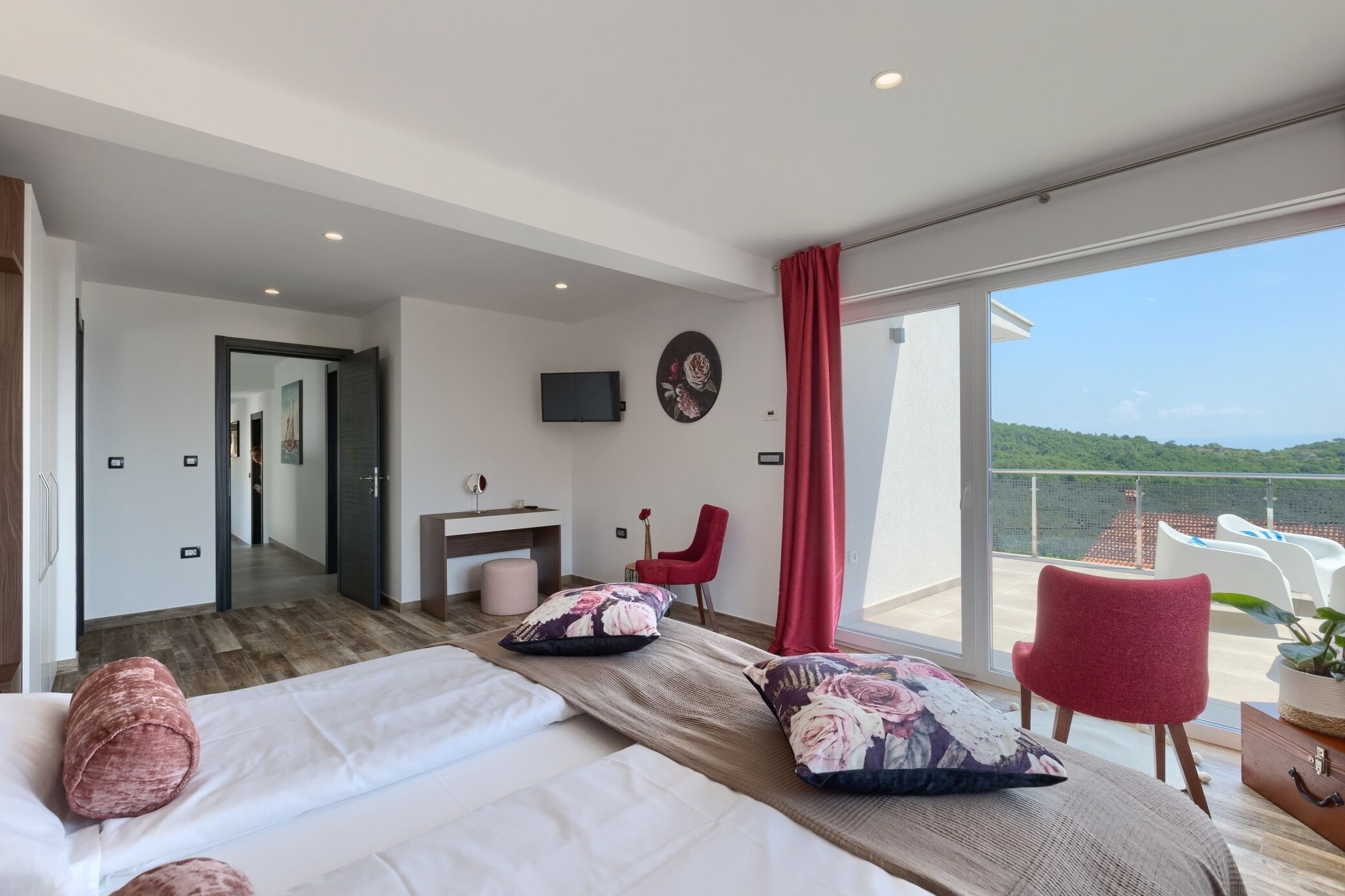 A modern hotel room with a balcony, a bed with floral pillows, a red chair, and a TV on the wall.