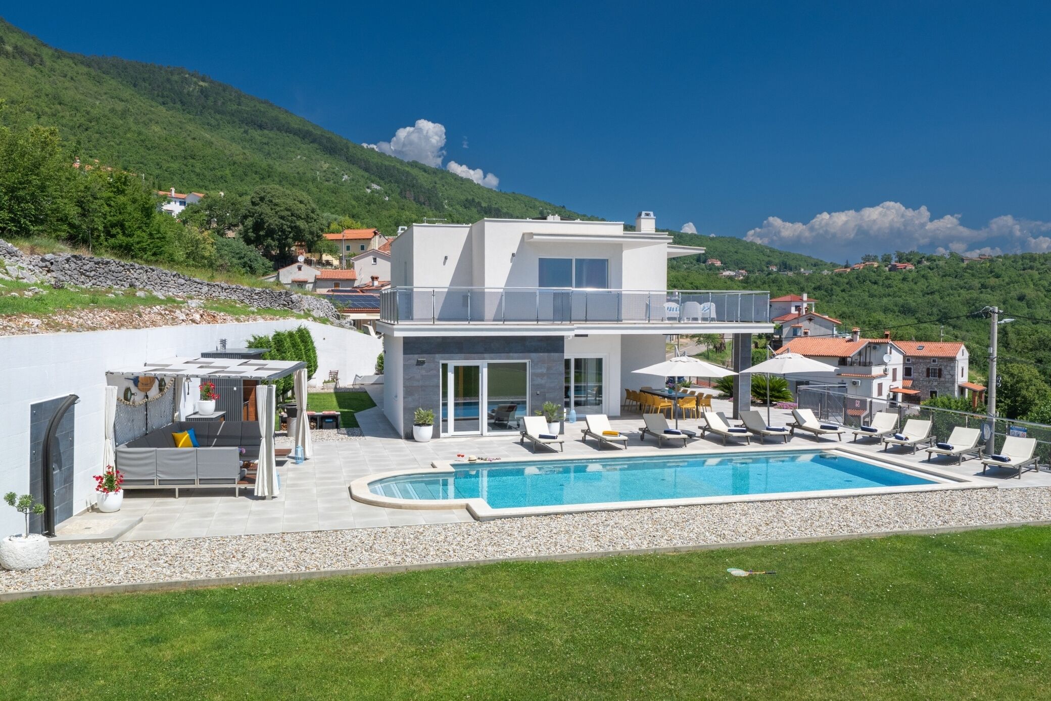 Une maison moderne à deux étages, dotée d’une piscine, d’un espace salon extérieur et offrant une vue sur les collines environnantes.