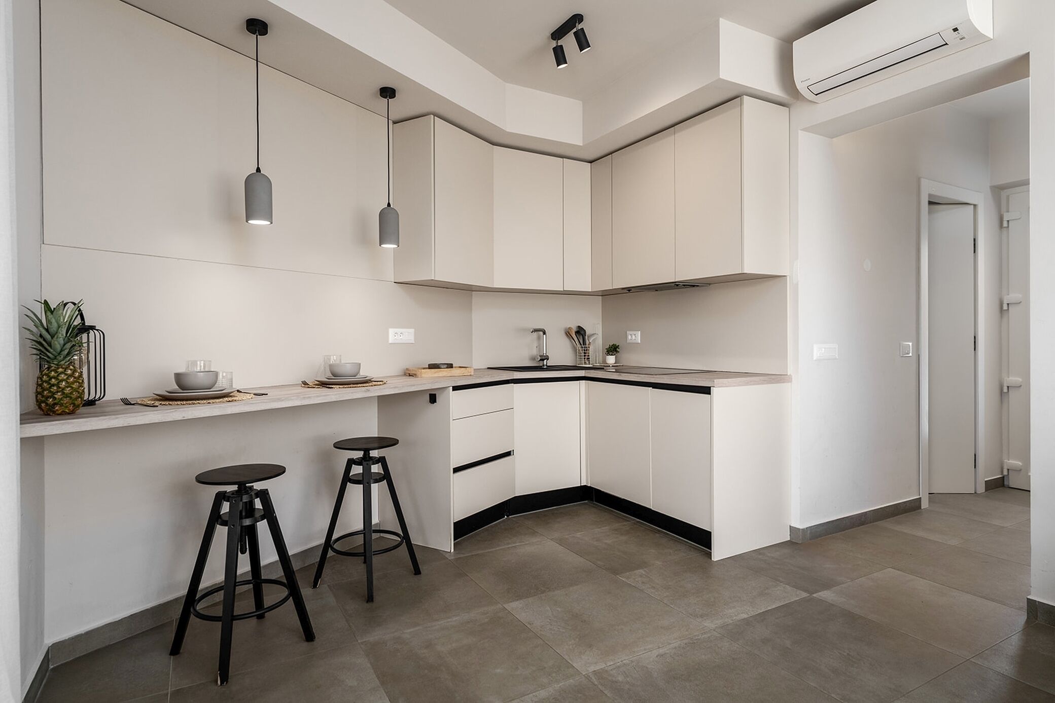 A modern kitchen with white cabinetry, a curved island, two bar stools, pendant lighting, and a visible air conditioning unit.