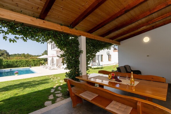 A wooden table with benches, a bottle of champagne, and a glass on a patio with a pool and greenery in the background.