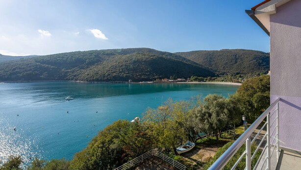 A balcony with a view of a turquoise lake, surrounded by greenery and hills.