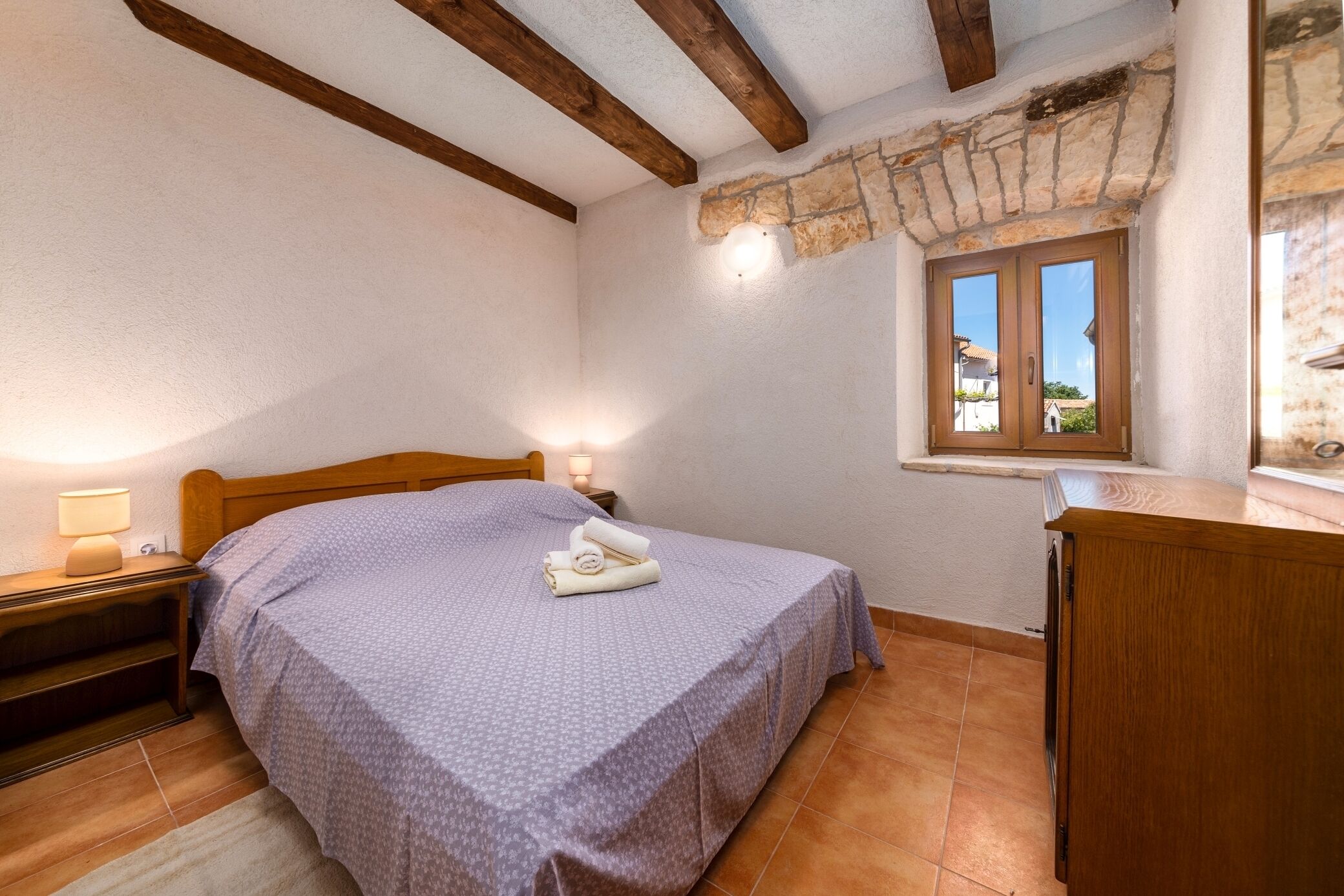 Une chambre  à coucher avec un lit, une tête de lit en bois, un mur en pierre, une fenêtre et une commode en bois.