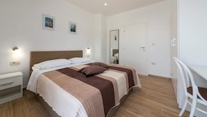 A bedroom with a large bed, a desk, and a chair. There are two wall-mounted lights and a door.